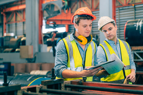 Two Caucasian industrial engineer men wear safety helmet vest inspecting checklist on clipboard. Worker partner discussing quality control audit in manufacturing factory. Metal coil background