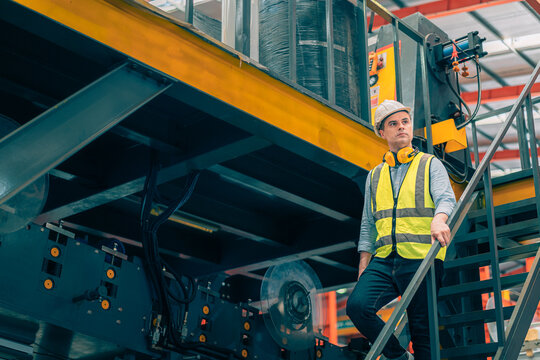 Caucasian industrial engineer man wear safety helmet vest walking down metal stairs in factory. Professional worker holding handrail safety routine. Heavy manufacturing plant background.