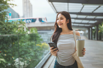 Happy asian woman using smartphone while holding reusable water bottle and carrying eco-friendly tote bag &ndash; sustainable lifestyle and conscious everyday choices in modern green city