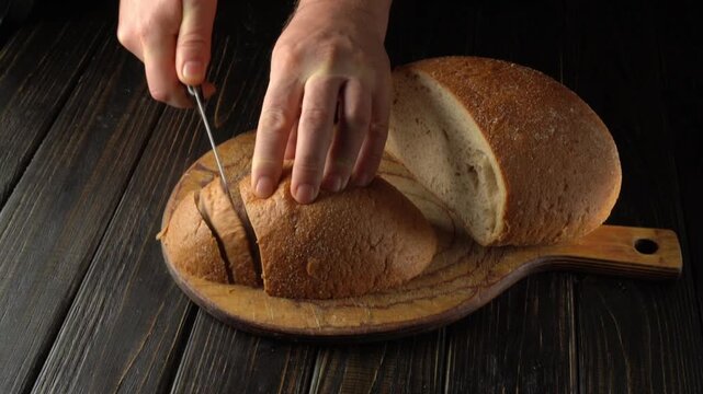Two hands hold a knife while slicing a loaf of bread on a wooden cutting board. The scene shows a kitchen with soft lighting