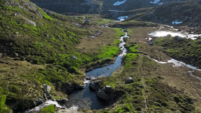 Aerial view of mountain stream along Kjerag hiking trail. Drone footage following a winding creek through alpine terrain and melting snowfields on a sunny summer day.