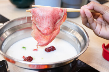 Hand using chopsticks to hold a large slice of raw marbled meat over a hot pot with milky white broth. Perfect for shabu-shabu or sukiyaki dining.