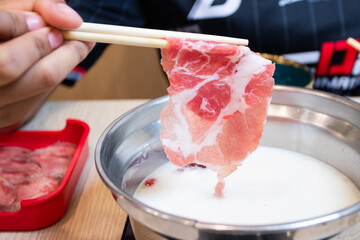 Hand using chopsticks to dip a slice of raw marbled meat into a hot pot with white broth. Perfect for shabu-shabu or sukiyaki dining.