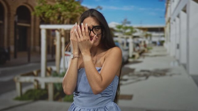 Woman covering face with hands on street wearing blue strapless dress and watch; embarrassment vulnerability.