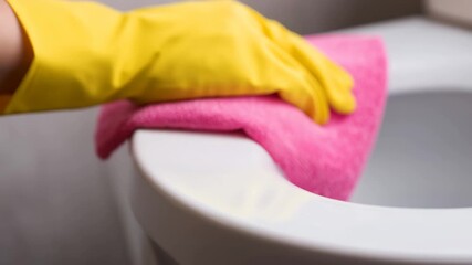 Yellow gloved hand wiping toilet rim with pink microfiber cloth close up hygiene cleaning bathroom sanitation routine domestic chores