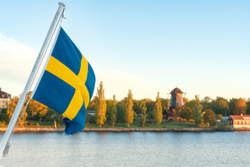Swedish flag waving over calm waters in the Stockholm archipelago, with Djurg&aring;rden island and autumn foliage in the background on a peaceful early October morning