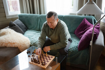 Mature man sitting on sofa and playing chess alone at home, relaxing and spending time in cozy living room interior. Home Leisure Activities