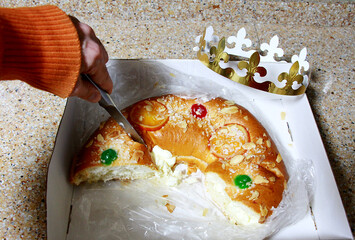 Rosc&oacute;n de reyes de Navidad relleno de nata y con fruta escarchada, az&uacute;car y corona de rey en caja con mano cortando trozo con cuchillo