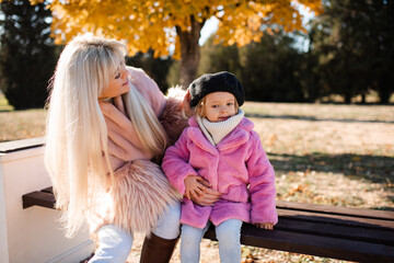 Fototapeta premium Smiling young adult woman holding child girl wearing fluffy coats over yellow leaves outdoors. Motherhood.