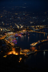 Fototapeta premium Night view of Alanya, Turkiye. Panoramic view of beach, city with street lamps, fortress walls, Red Tower, and Mediterranean Sea. Tourism and seaside recreation