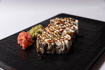 Sushi rolls topped with creamy mayonnaise and teriyaki sauce, served on a black plate with wasabi and pickled ginger