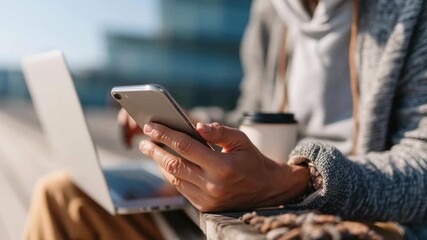 A man is sitting on a bench with a laptop and a cell phone in his hands. He is looking at his cell phone while holding a cup of coffee. The scene suggests that he might be multitasking