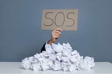 Businessman holding sign with word SOS at white table with pile of crumpled paper balls against...