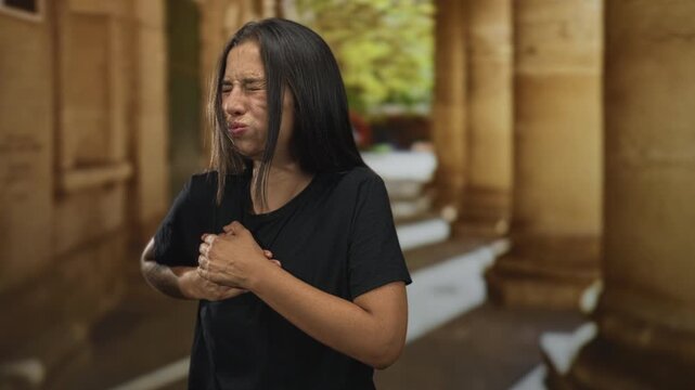 Young hispanic woman clutching chest for heartattack in old town building corridor, hands pressing sternum and eyes closed in grimace; panic.