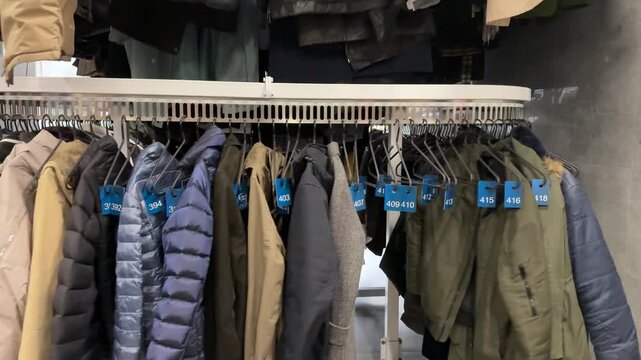 Rows Of Winter Coats Hanging, Shop Owner Sorting Preloved Inventory Beneath Crowded Rack, Mixed Textures Wool Denim Leather, Muted And Bright Colors, Cozy Cluttered Atmosphere, Sustainable