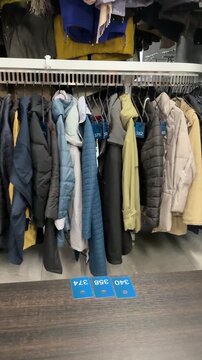 Rows Of Winter Coats Hanging, Shop Owner Sorting Preloved Inventory Beneath Crowded Rack, Mixed Textures Wool Denim Leather, Muted And Bright Colors, Cozy Cluttered Atmosphere, Sustainable