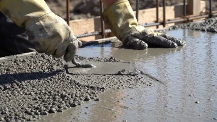 Craftsman with gloves meticulously shapes wet concrete on a bustling construction site under bright sunlight, emphasizing artistry and precision in each stroke of the trowel.
