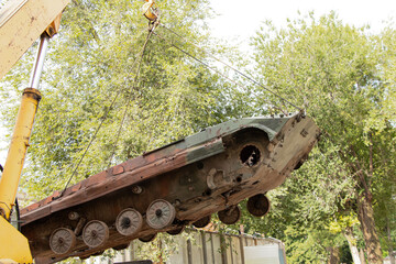 Moving decommissioned military equipment with a crane. A rusty, crewless armored vehicle is lifted for transport against a backdrop of trees, daylight in Dnipro