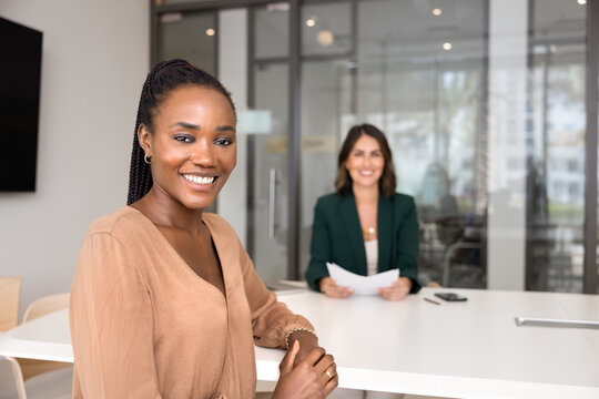 Positive beautiful young African American job candidate posing for portrait at table of recruiter on interview, looking at camera, smiling, sitting at meeting desk with HR manager in burred background