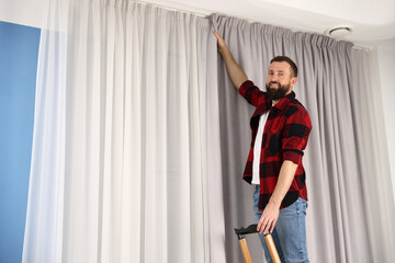 Man with ladder hanging up window curtains at home, low angle view. Space for text © New Africa