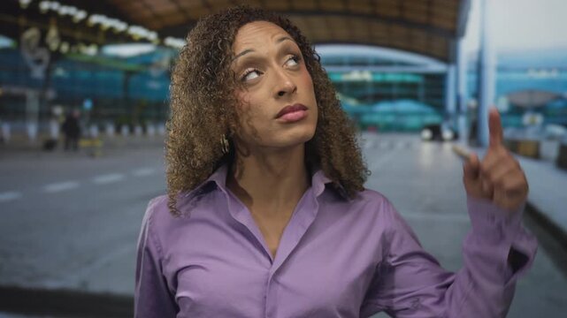 Curly haired transgender woman in purple shirt giving thumbs down gesture at airport terminal walkway; disapproval.