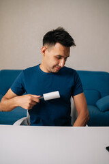 Vertical portrait of smiling man sitting at white table using sticky lint roller to remove dust, hair and lint from blue t-shirt, looking pleased with cleaning results in modern apartment.
