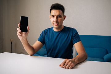 Portrait of handsome young adult man looking at camera displaying smartphone with blank screen, creating ideal mockup for advertisements or app promotions in modern workspace, sitting at white table,