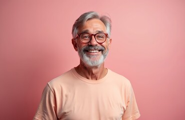 Obraz premium Elderly man with grey hair and beard smiles wearing glasses and peach t-shirt. He has a cheerful expression. This studio portrait shot against a pink background.