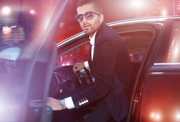 A handsome man in a suit gets into a car in the glow of night headlights.