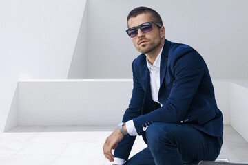 A man in a dark blue suit sitting against a white wall. The design of a representative brand in minimalistic interiors.