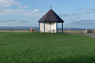 A white gazebo with a dark roof sits on green grass overlooking a blue sea under a bright, clear sky, creating a peaceful scene in Llanfairfechan - Wales - UK