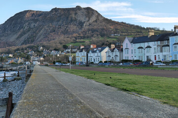 Coastal townscape: pastel houses line a green verge, a grey promenade leads to distant buildings, dominated by a large, rugged mountain under soft, warm light in Llanfairfechan - Wales - UK