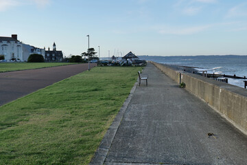 Seaside promenade: concrete walkway, grassy verge, bench, sea wall, distant buildings, calm blue sea, clear sky, bright daylight, peaceful atmosphere, horizontal composition in Llanfairfechan - Wales 