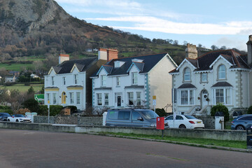 Row of white houses with bay windows lines a street, cars parked, against a backdrop of a large, brown, mountainous hill under daylight in Llanfairfechan - Wales - UK