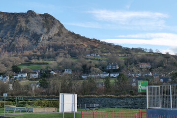 A rocky mountain looms over a village nestled on its slope, under a bright blue sky, with a green field in the foreground in Llanfairfechan - Wales - UK