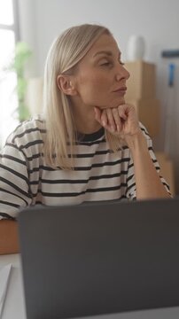 Woman resting hand on chin at home office desk with laptop and stacked moving boxes; calm reflection.