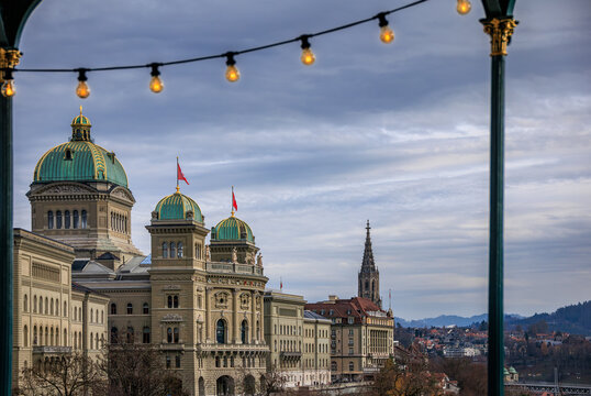 A string of warm Christmas lights of the Christmas Market frame a winter view of the Federal Palace and Minster spire at twilight in Bern, Switzerland