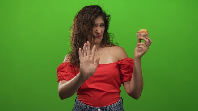 Woman wearing red off shoulder top holding muffin with raised hand and disgusted face in studio with green backdrop; disgust aversion refusal hesitation.