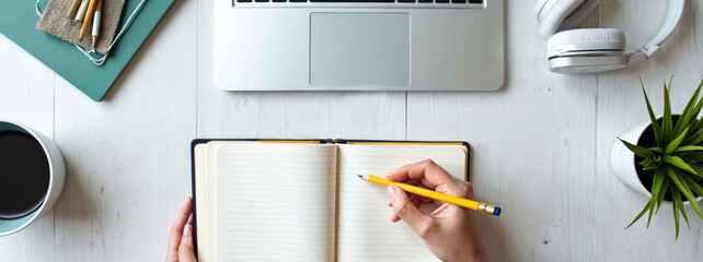 Person writes in notebook next to laptop and headphones. Coffee cup and plant on desk. Remote work or study from home setup, planning or note taking.