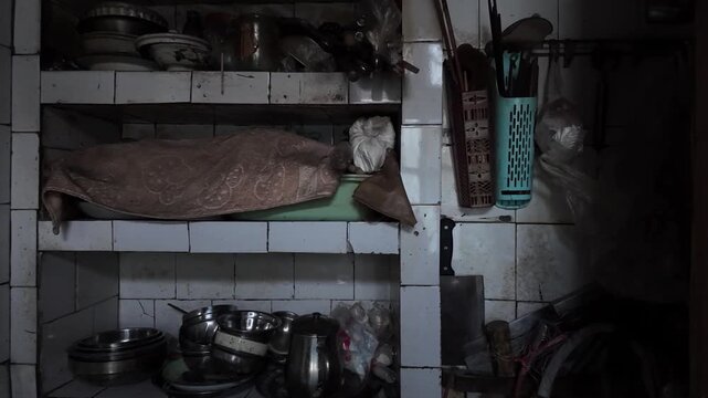 An old, cluttered kitchen storage area with open tiled shelves holding dusty cooking utensils. Household items convey a sense of aging, third-world domestic space, disorganized, poor living conditions