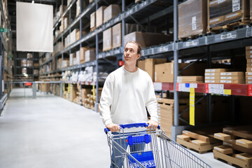 Young man pushing a shopping cart in a large furniture warehouse store, looking up at tall shelves filled with boxes. © Kyrylo