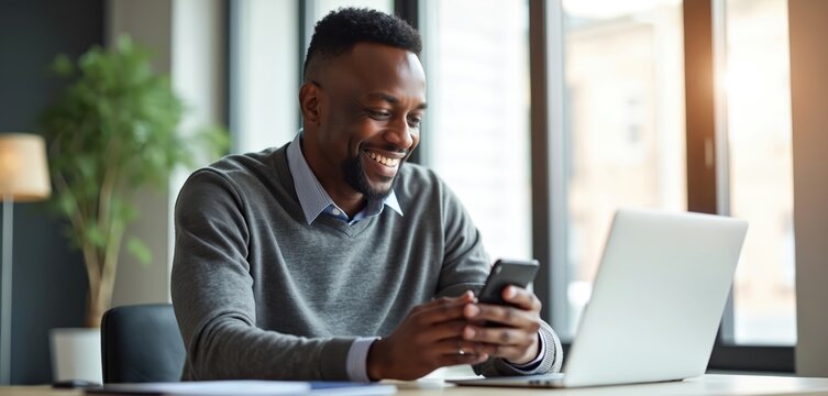 Smiling African American man types on phone at modern office desk. Laptop open, he browses internet, chats, enjoys workday. Natural light fills room. - Powered by Adobe
