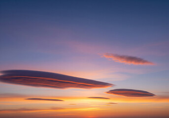 Vibrant sunset hues paint the expansive sky, illuminating a captivating formation of distinct lenticular clouds, creating a serene and breathtaking natural spectacle that inspires awe