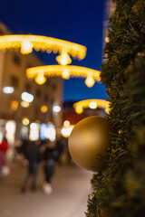 Close-up of a golden Christmas ornament on a decorated evergreen tree with warm festive lights and city street bokeh in the background at dusk. Cozy holiday atmosphere, winter celebration, urban Chris