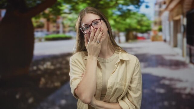 Young blonde woman wearing glasses and a light shirt covers mouth with hand on bustling city street; amusement.