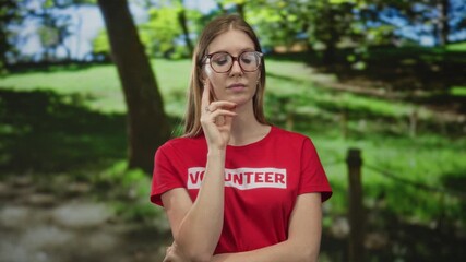 Young blonde volunteer woman wearing red shirt with hand on chin gesture in sunny green park; contemplation.