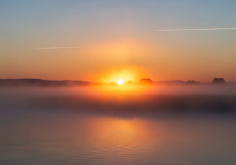 Golden sunrise through morning mist over serene lake, reflecting warm light on calm waters, with distant forest, capturing environmental beauty and tranquil dawn