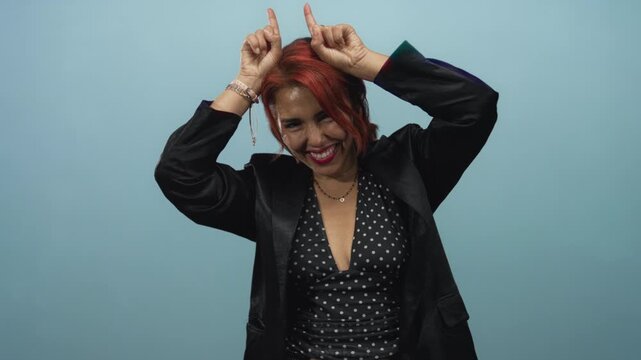 Woman with both index fingers raised near head like horns, wearing polka dot top and black blazer in light blue studio; playful confidence.