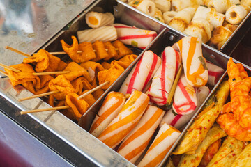 A variety of colorful Korean street food snacks, including fish cake, fish balls, surimi, and chikuwa, are neatly organized in metal compartments at a food stall.