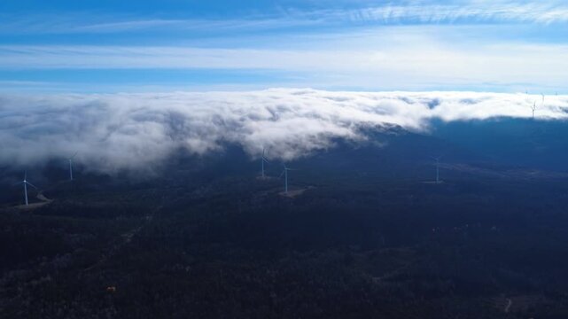 High-altitude aerial view above a blanket of low clouds covering mountains dotted with wind turbines under a blue sky. Matapedia Valley, Quebec, Camada, 2025.